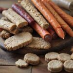 A hyperrealistic close-up of various healthy, natural dog treats scattered on a rustic wooden table. Include carrot sticks, baked biscuits, and dried meat pieces. Soft, natural lighting highlights the textures, with no text.