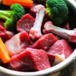 Hunde BARF Plan erstellen: Schritt für Schritt zur artgerechten Rohfütterung! Extreme close-up, hyperrealistic image of a stainless steel dog bowl brimming with fresh raw beef cuts, chicken bones, bright orange carrots, and vivid green broccoli florets. Condensation on the bowl, soft natural daylight from a kitchen window, shallow depth of field, sharp focus on food textures. Photorealistic, no text.