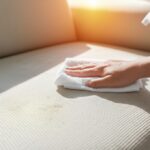 Hyperrealistic close-up of a hand cleaning a dog urine stain on a beige fabric sofa with a cloth in a sunlit living room. Enzyme cleaner spray bottle visible. Focus on fabric texture and stain detail. Shallow depth of field. Photorealistic, no text.